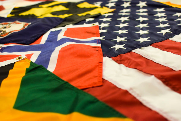 Layered display of U.S., state, and world flags at Kengla Flag Co., showing varied colors, emblems, and proportions.